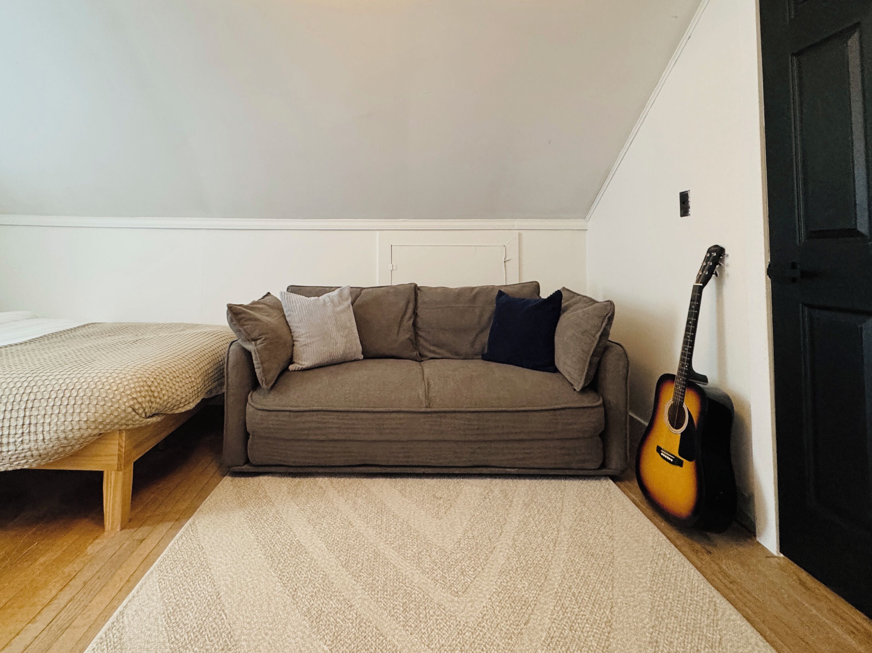 Ein kleiner, gemütlicher Raum mit einem braunen Sofa, einem hölzernen Bett und einer an der Wand angelehnten Gitarre.