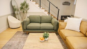 A living room with a green sofa, a yellow sofa, a coffee table with a bowl of fruit and a plant, a white chair, and a staircase in the background. - 2 Min Walk to Windham Town | Hot Tub | Fire Pit (Windham)