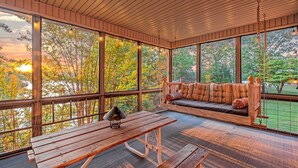 Una terraza de madera con un banco y una mesa, ventanas amplias y vista a árboles.