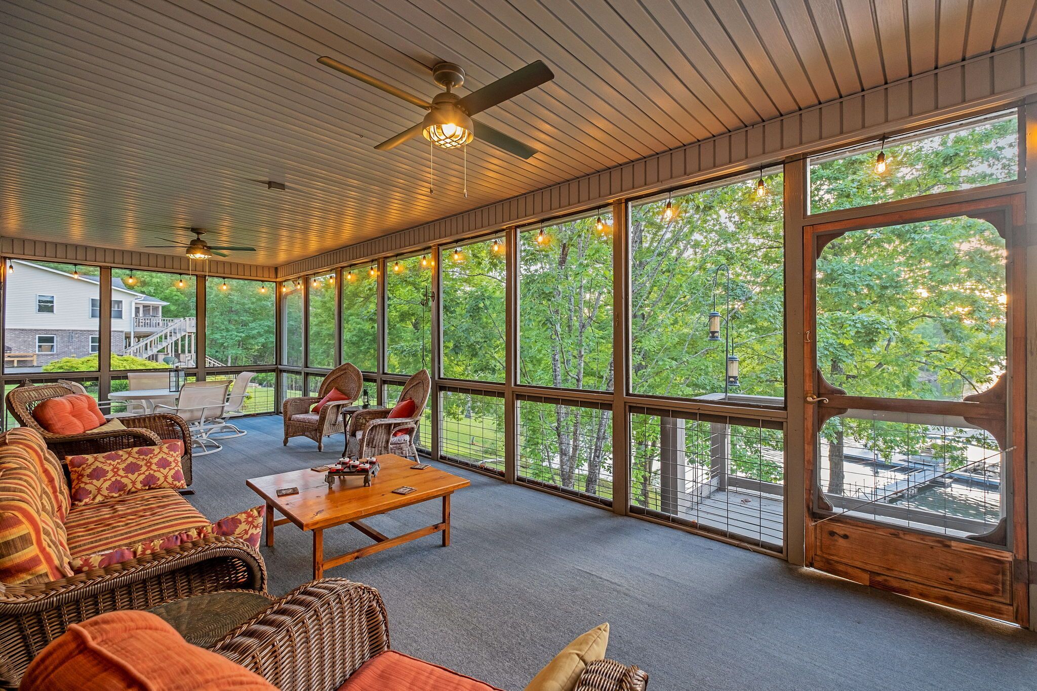 A covered outdoor seating area with wicker furniture, a wooden table, and large windows offering a view of trees and a house.