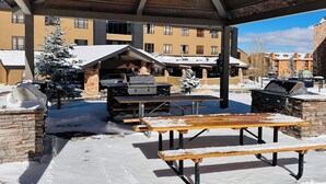 A covered outdoor area with picnic tables and barbecue grills, surrounded by buildings with snow on the roofs. - Resort Condo NEXT to Ski Run w/ Pool & Hot tub! (Brian Head)