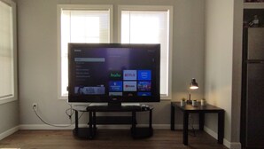 Een flatscreen televisie op een standaard in een kamer met een raam, een kleine tafel met een lamp en een koelkast.