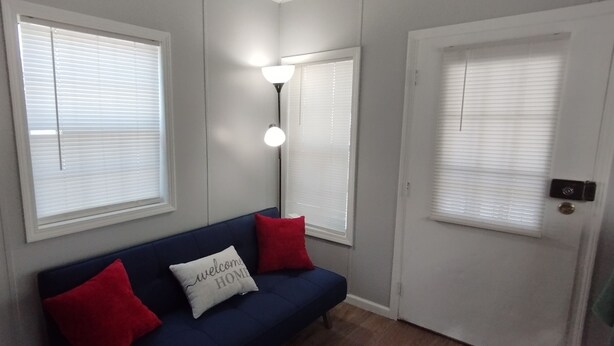 Een kamer met een blauwe bank, twee rode kussens, een wit kussen met de tekst 'welcome home', een wandlamp en een deur met een raam.