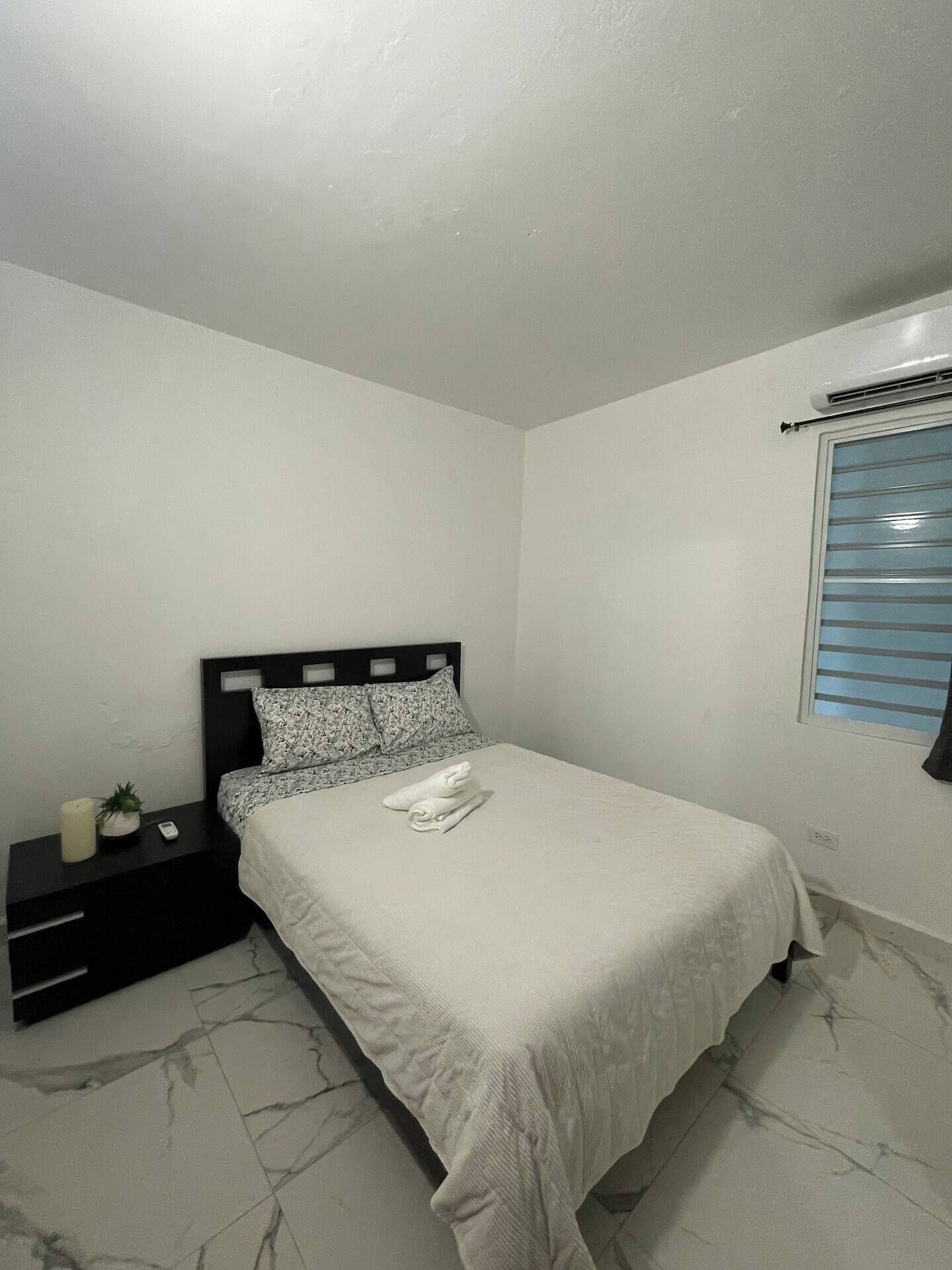 A bedroom with a bed, a nightstand, a wall-mounted air conditioner, and a window with blinds.
