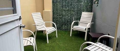 Un petit espace patio avec du gazon synthétique, deux chaises en métal blanches et un mur décoratif orné de plantes en suspension.
