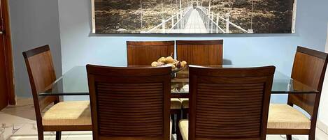 Un espace repas doté d’un sol en marbre, de chaises en bois avec des sièges rembourrés, d’une table à plateau en verre et d’un grand tableau représentant un pont, accroché au mur.