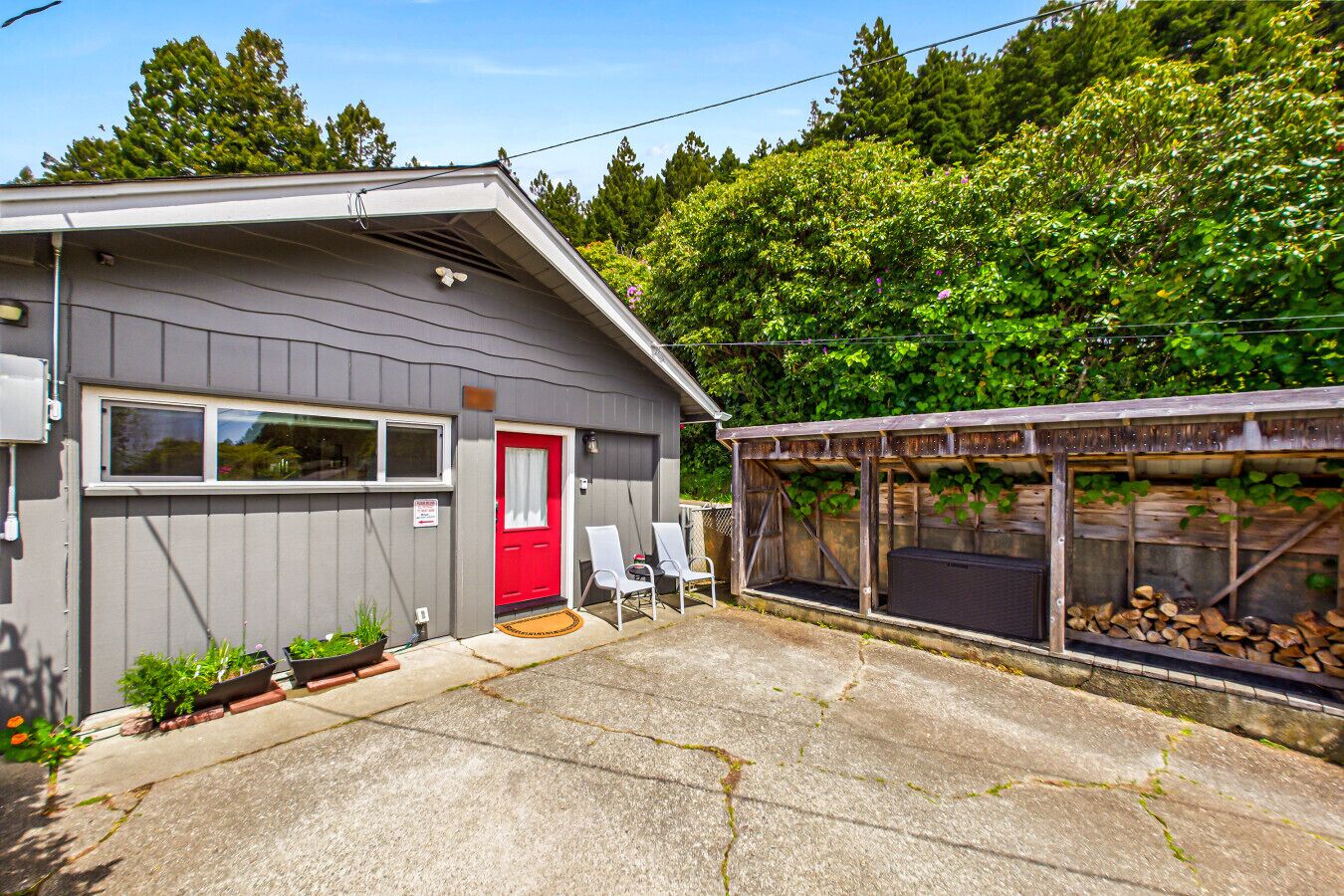 A small, single-story building with a red door, a covered outdoor area with chairs, and a stack of firewood.