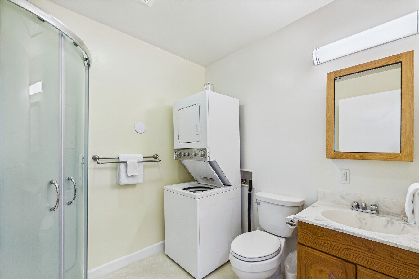 A bathroom with a glass shower enclosure, a washing machine, a toilet, and a vanity with a mirror.