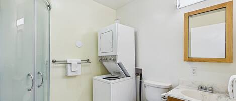 A bathroom with a glass shower enclosure, a washing machine, a toilet, and a vanity with a mirror.