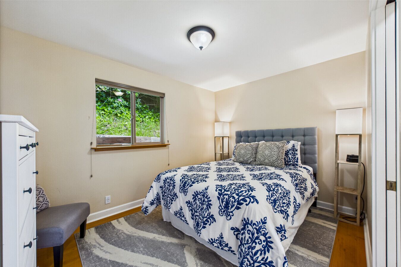 Une chambre à coucher avec un lit, une commode, un banc, une fenêtre et une lampe.