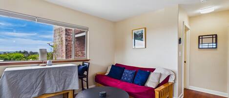 Una habitación con una mesa de comedor, un sofá rojo, una pequeña mesa redonda y vista a un edificio de ladrillo a través de una ventana.