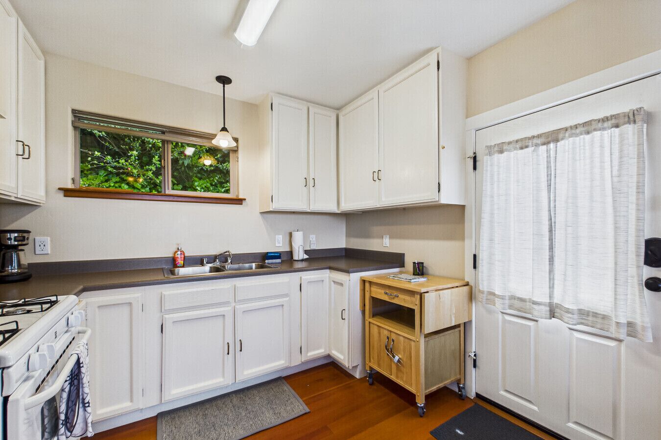 A compact kitchen with white cabinets, a built-in stove, a small wooden table, and a window with a view of greenery.