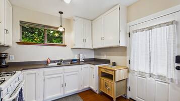 A compact kitchen with white cabinets, a built-in stove, a small wooden table, and a window with a view of greenery.