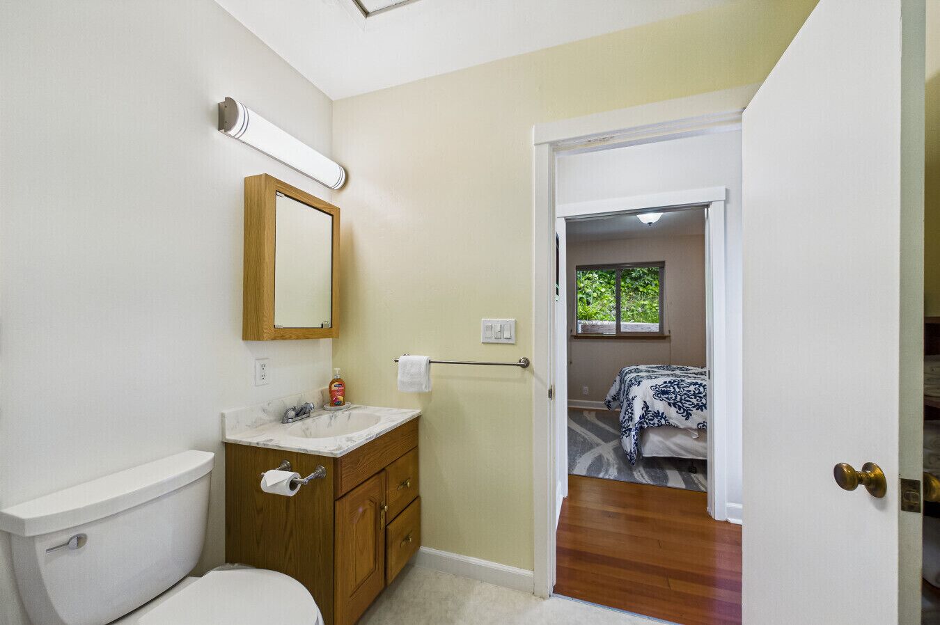 A compact bathroom with a toilet, sink, and mirror. A towel rack is visible, and a window provides natural light to a bedroom with a bed and patterned bedding.