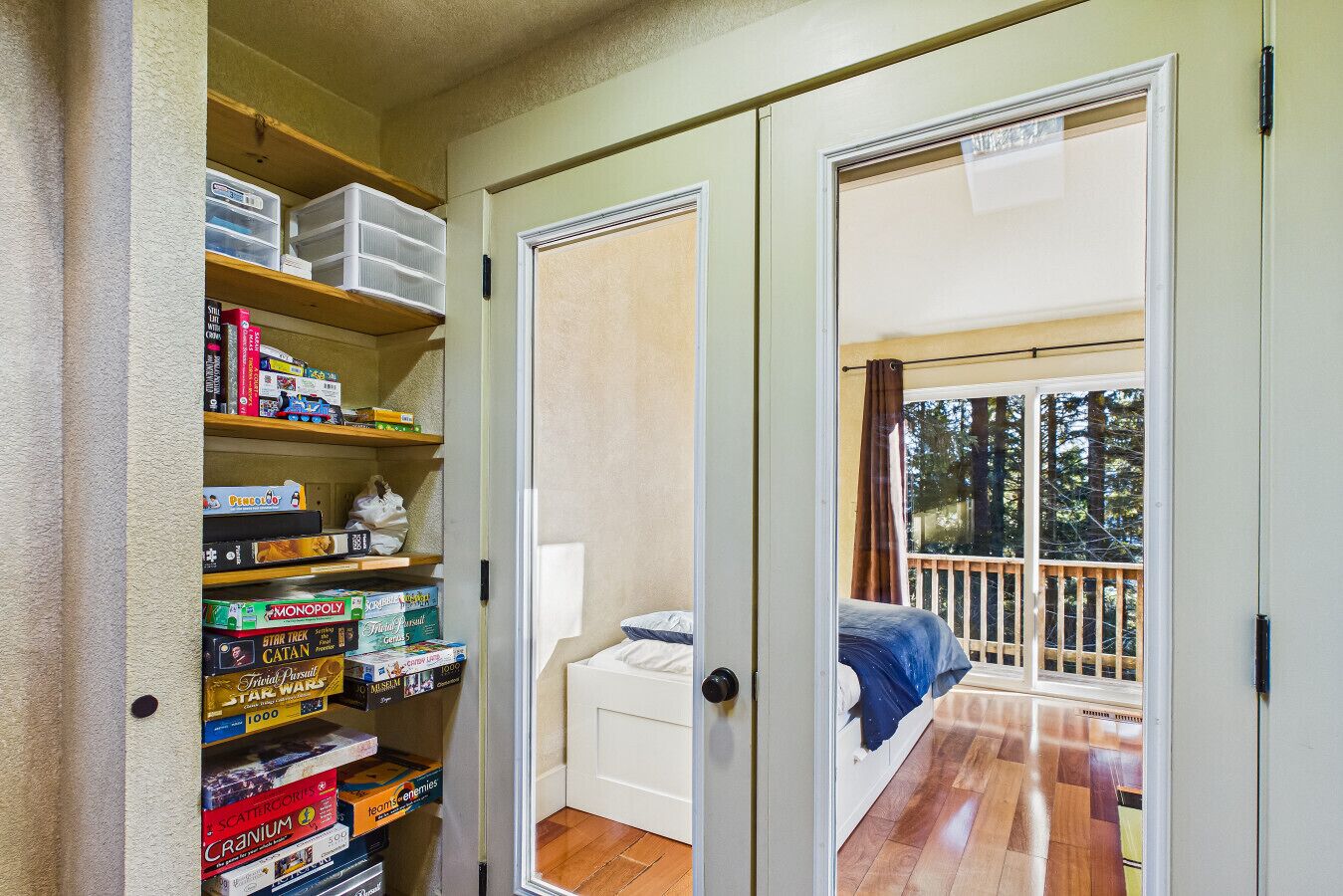 Une chambre avec un lit, un placard contenant des étagères remplies de jeux de société, et un balcon doté d’une terrasse en bois et d’une rambarde.