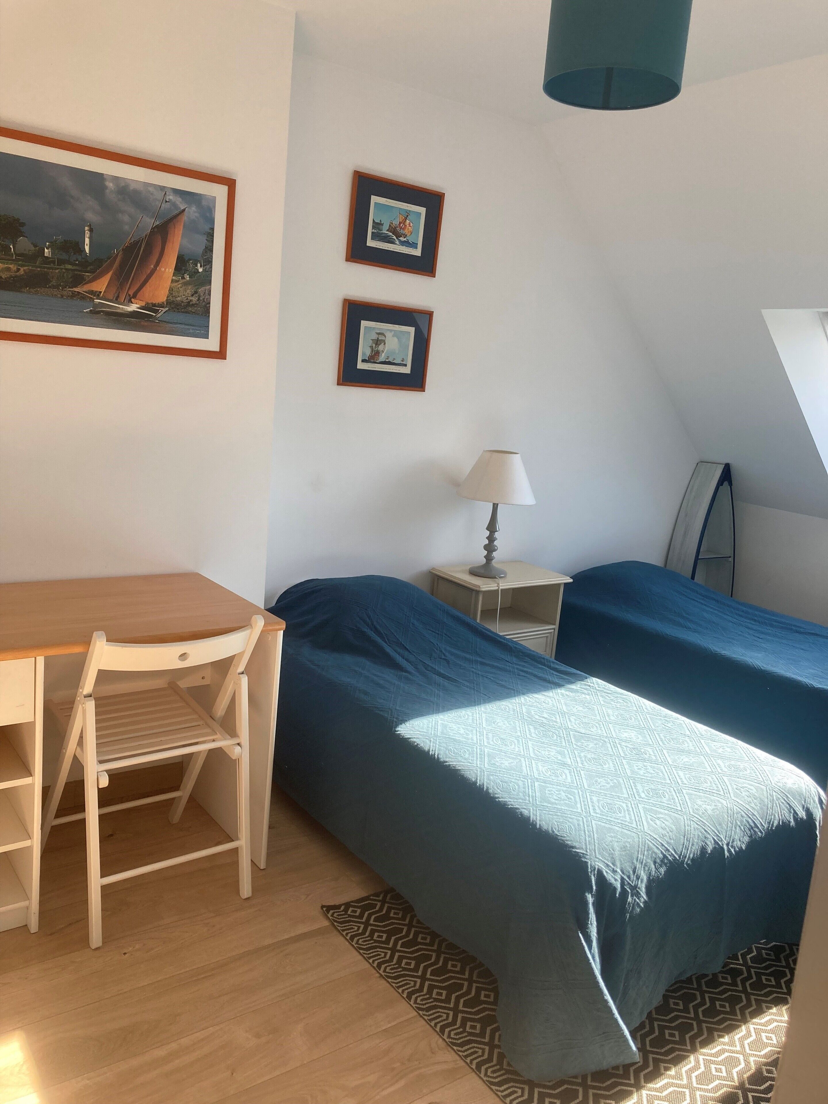 A room with a desk, two beds, a chair, a lamp, and framed pictures on the wall.