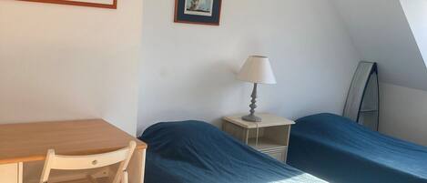 A room with a desk, two beds, a chair, a lamp, and framed pictures on the wall.