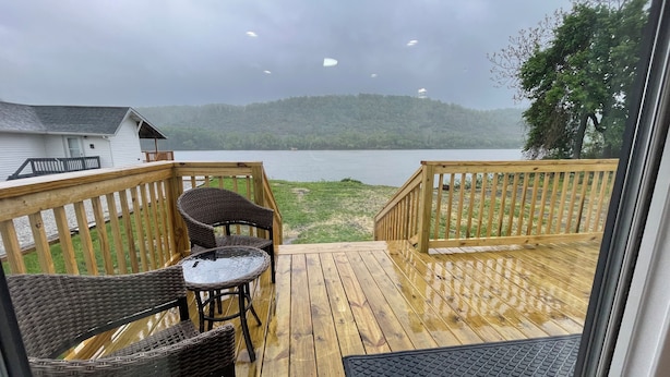 Une terrasse en bois, meublée de meubles en osier et dotée d’une table en verre, qui offre une vue sur un lac.