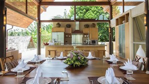 A dining area with a wooden table set for a meal, surrounded by wooden chairs, with a view of the kitchen and outdoor greenery.