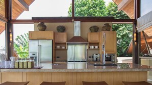 A modern kitchen with wooden cabinets, a stainless steel refrigerator, a coffee machine, and a large window overlooking greenery.