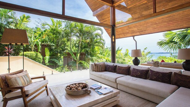 A spacious living area with a large window overlooking lush greenery and a view of the sea. There is a sofa, a coffee table with a bowl of nuts, and a chair.