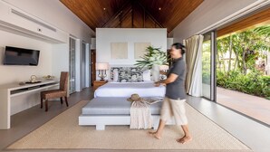 A modern bedroom with a large bed, a desk, a TV, and a view of a tropical garden.