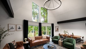 A spacious living room with a brown leather sofa, green armchairs, a round coffee table, and a dining area with a wooden table and chairs. Large windows provide natural light and a view of trees. - Bourbon Trail*River Views*Hot Tub*Sauna*EVSE*WiFi (Frankfort)