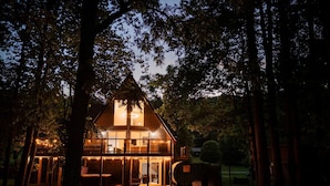 A two-story house with a glass facade, surrounded by trees, and illuminated by string lights. - Bourbon Trail*River Views*Hot Tub*Sauna*EVSE*WiFi (Frankfort)