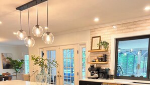 A modern kitchen with a large marble island, black bar stools, pendant lights, and a view of the outdoors through a large window.