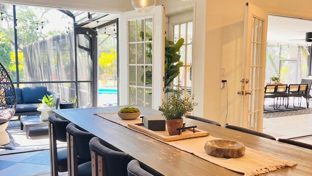 A modern kitchen with a long wooden table, black chairs, hanging pendant lights, and large windows offering views of the outdoors.