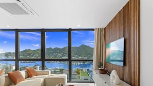 A modern living room with a large TV, a sofa, a coffee table, and a view of the sea and mountains. - B231 - Serene Chic 2 Bedroom (Sint Maarten)