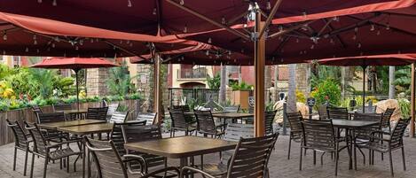 An outdoor patio with tables and chairs under red umbrellas, surrounded by greenery and a stone wall.