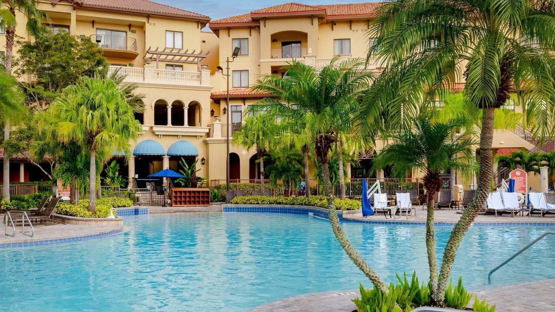 Un hotel con piscina, palmeras y una zona cubierta con sombrillas.