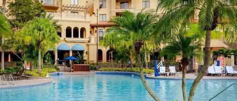 Un hotel con piscina, palmeras y una zona cubierta con sombrillas.