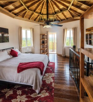 Een slaapkamer met een houten plafond, een bed met een rode plaid, een houten vloer, een balkon met een hekwerk en uitzicht op de omgeving.