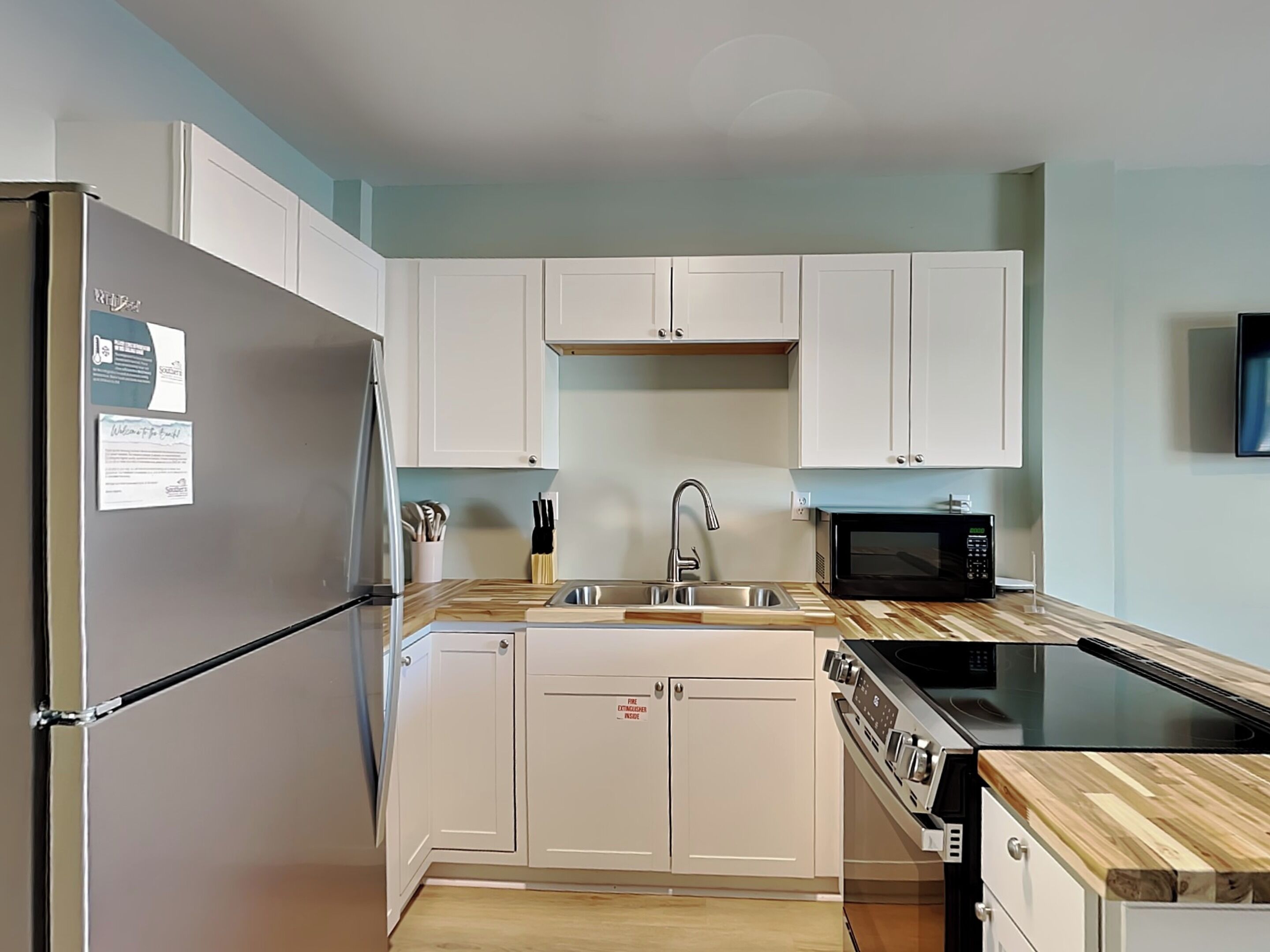 A modern kitchen with white cabinets, a stainless steel refrigerator, a microwave, a sink, and a stove.