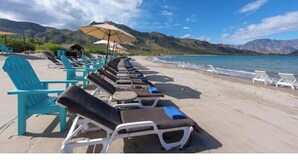 Una playa con filas de sillas de playa y sombrillas, vista despejada al mar y montañas al fondo.