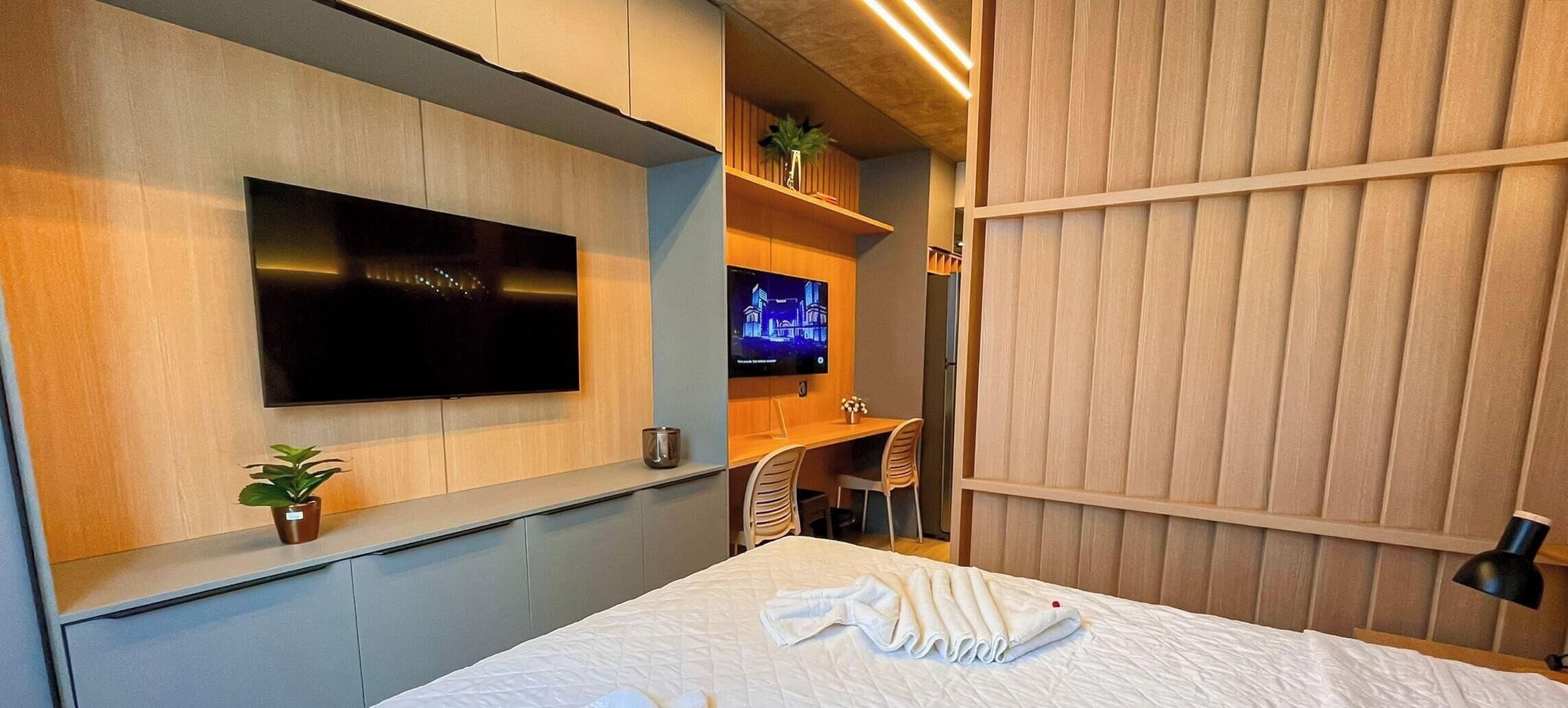 Quarto de hotel com uma cama, televisão na parede, escrivaninha com cadeira e uma planta.