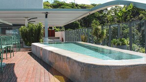 A rectangular swimming pool with a stone edge, surrounded by a metal fence and potted plants. There are green chairs and a covered area with a roof. - Setai Yacht Premium II - By Almare Flats (João Pessoa)