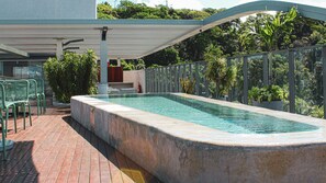 Una piscina rectangular con borde de piedra, rodeada por una valla metálica y plantas en macetas. Hay sillas verdes y una zona cubierta con techo.