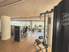 A gym with cardio machines, weightlifting equipment, and a view of palm trees through large windows. - Setai Yacht Premium II - By Almare Flats (João Pessoa)