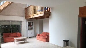 Un vaste espace de vie comprenant deux canapés rouges, une table basse blanche et un banc en bois. Un meuble blanc rempli de livres est adossé au mur, et une poubelle est visible.
