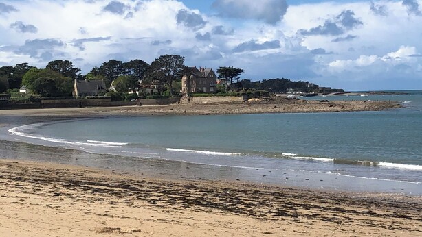 Un paysage côtier avec une plage de sable, une mer calme et une rangée de maisons surmontées d’une tour imposante.