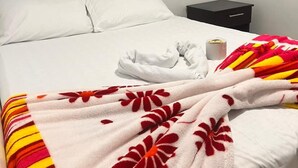 Un lit avec du linge de lit blanc, une couverture à motifs floraux, une petite table de chevet avec une lampe et une commode.