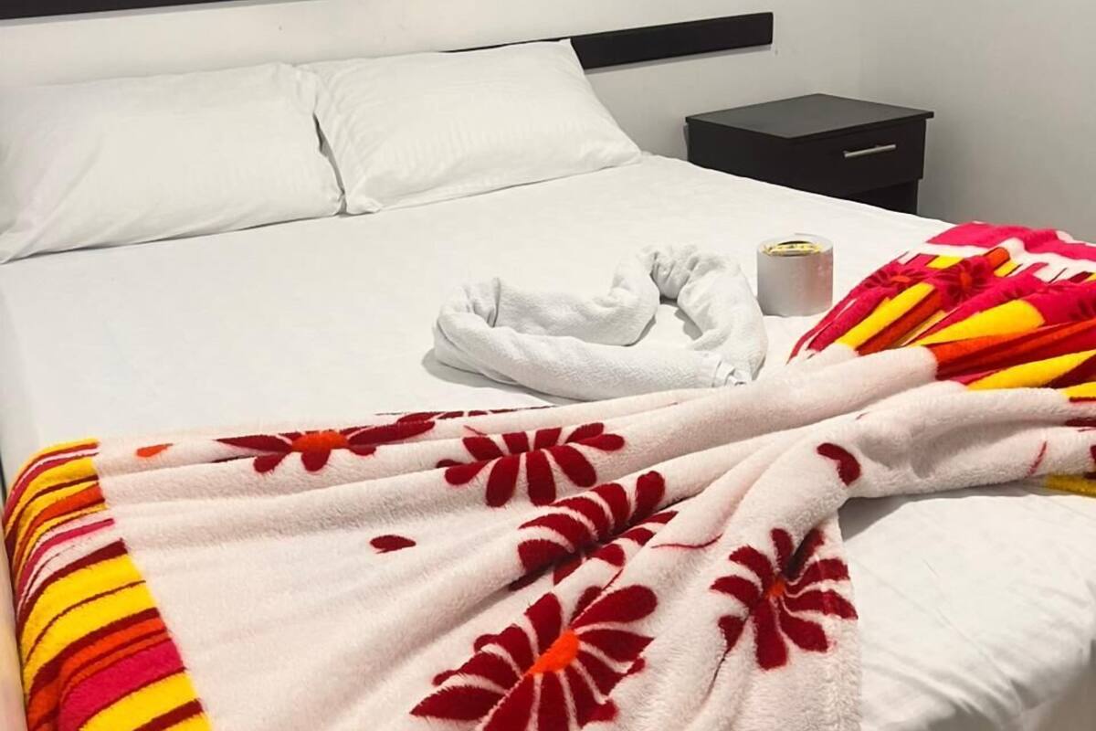 Una cama con sábanas blancas, una colcha floral, una mesita de noche pequeña con una lámpara y otra mesita de noche.