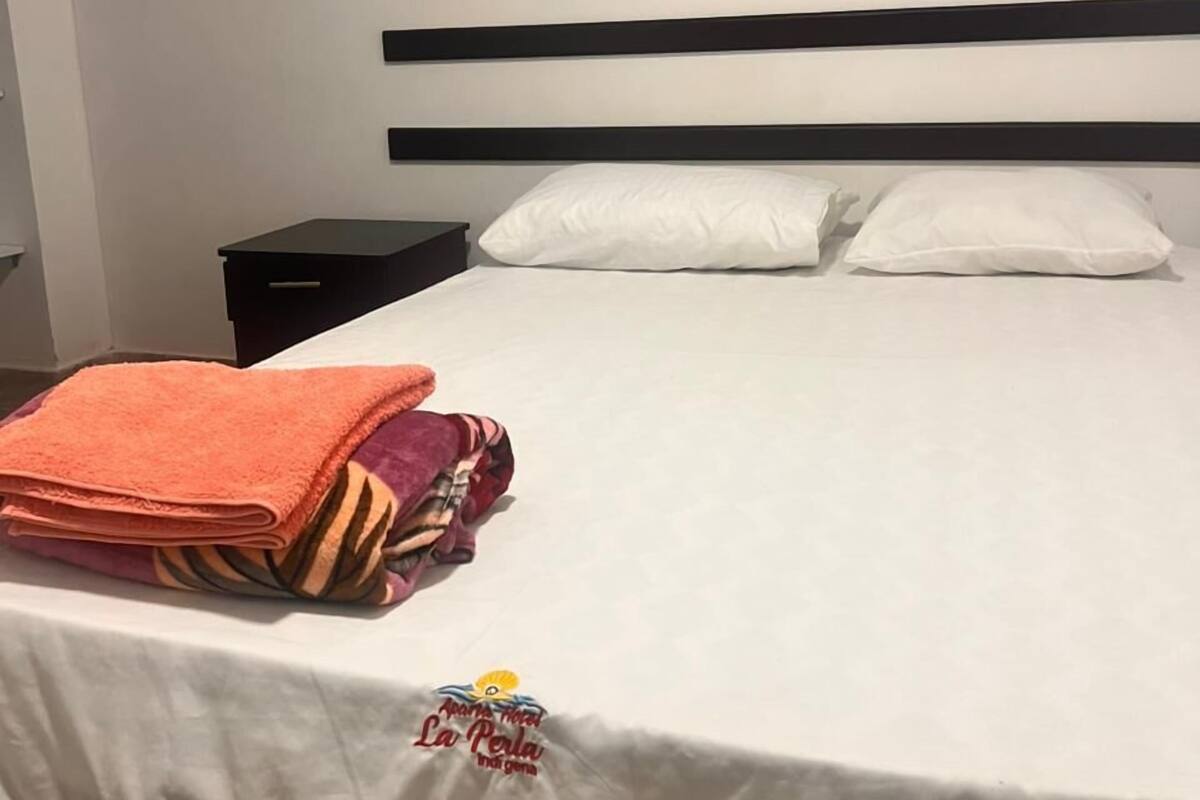 Una cama con sábanas blancas, cabecera negra y una toalla naranja doblada con cuidado al lado.