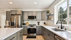 A modern kitchen with grey cabinets, stainless steel appliances, a large window, and a black rug. - Pelican's Rock: 10 min walk to downtown, beach & park + Deck + Fire pit (Sturgeon Bay)