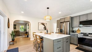 A modern kitchen with a central island, grey cabinetry, stainless steel appliances, and a dining area with chairs and a table. - Pelican's Rock: 10 min walk to downtown, beach & park + Deck + Fire pit (Sturgeon Bay)