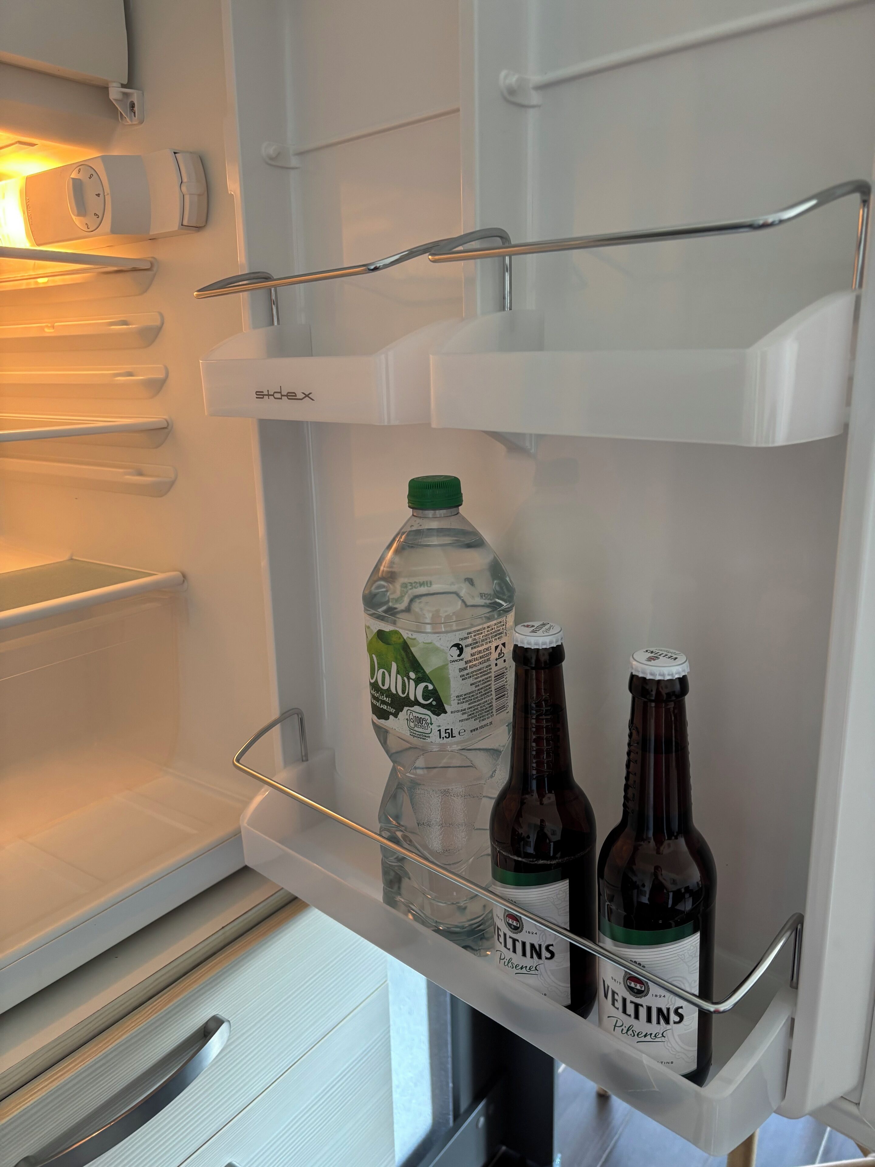 Ein Kühlschrank mit einer Ablage, auf der eine Wasserflasche, zwei Bierflaschen und eine Cola stehen.