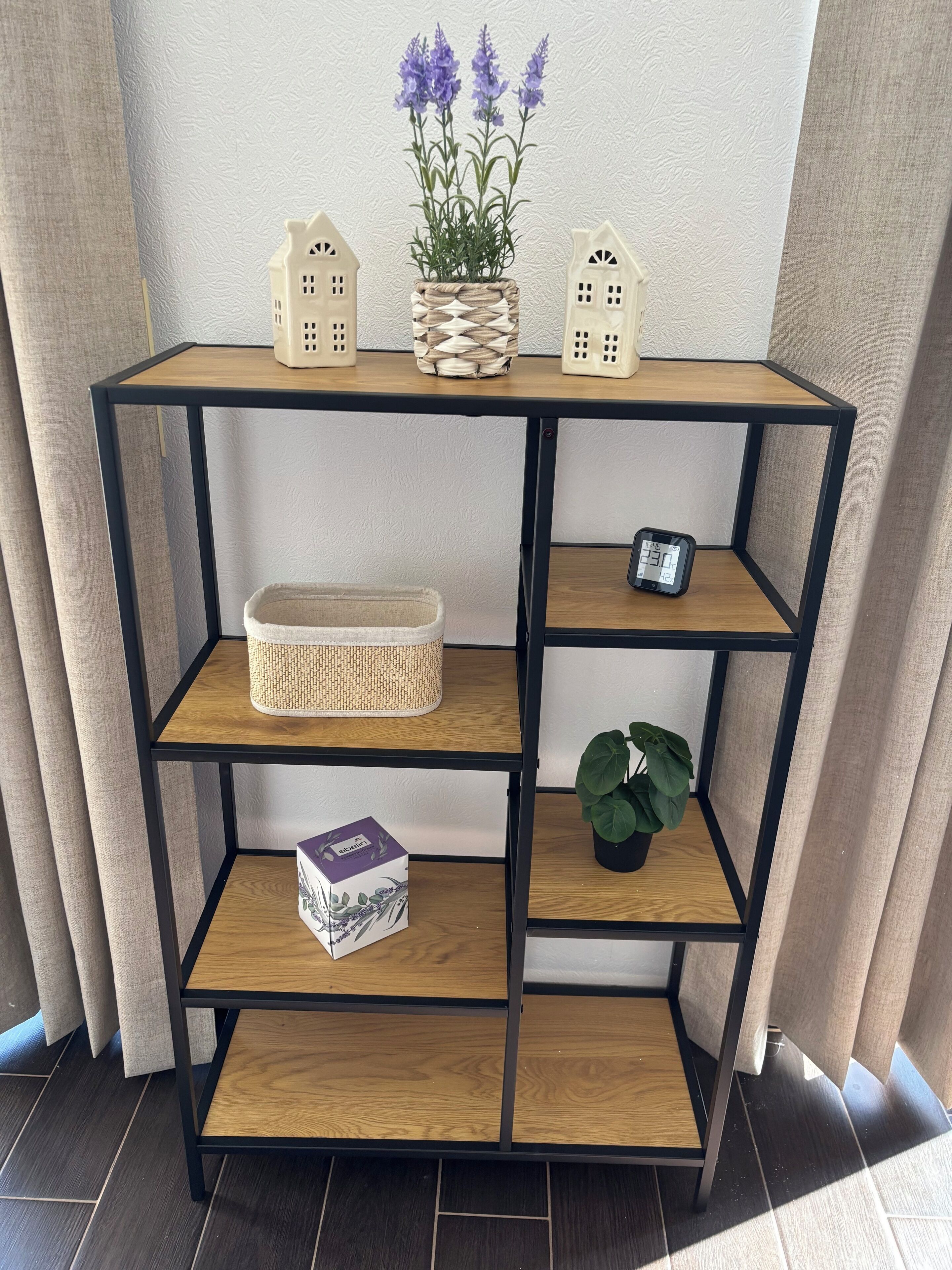Een meerlaagse houten plankenwand met een gevlochten pot bovenop, een lamp in de vorm van een huisje, en een plant in een pot.